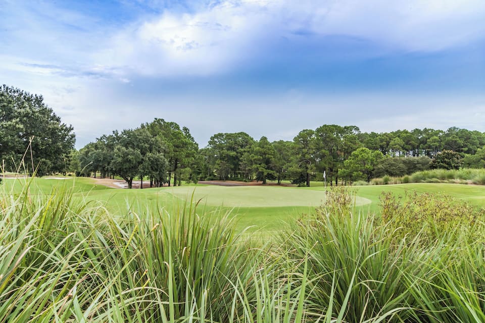 Ein Golfplatz mit üppigem Grün, hohem Gras im Vordergrund und einer Baumreihe im Hintergrund unter teils bewölktem Himmel.