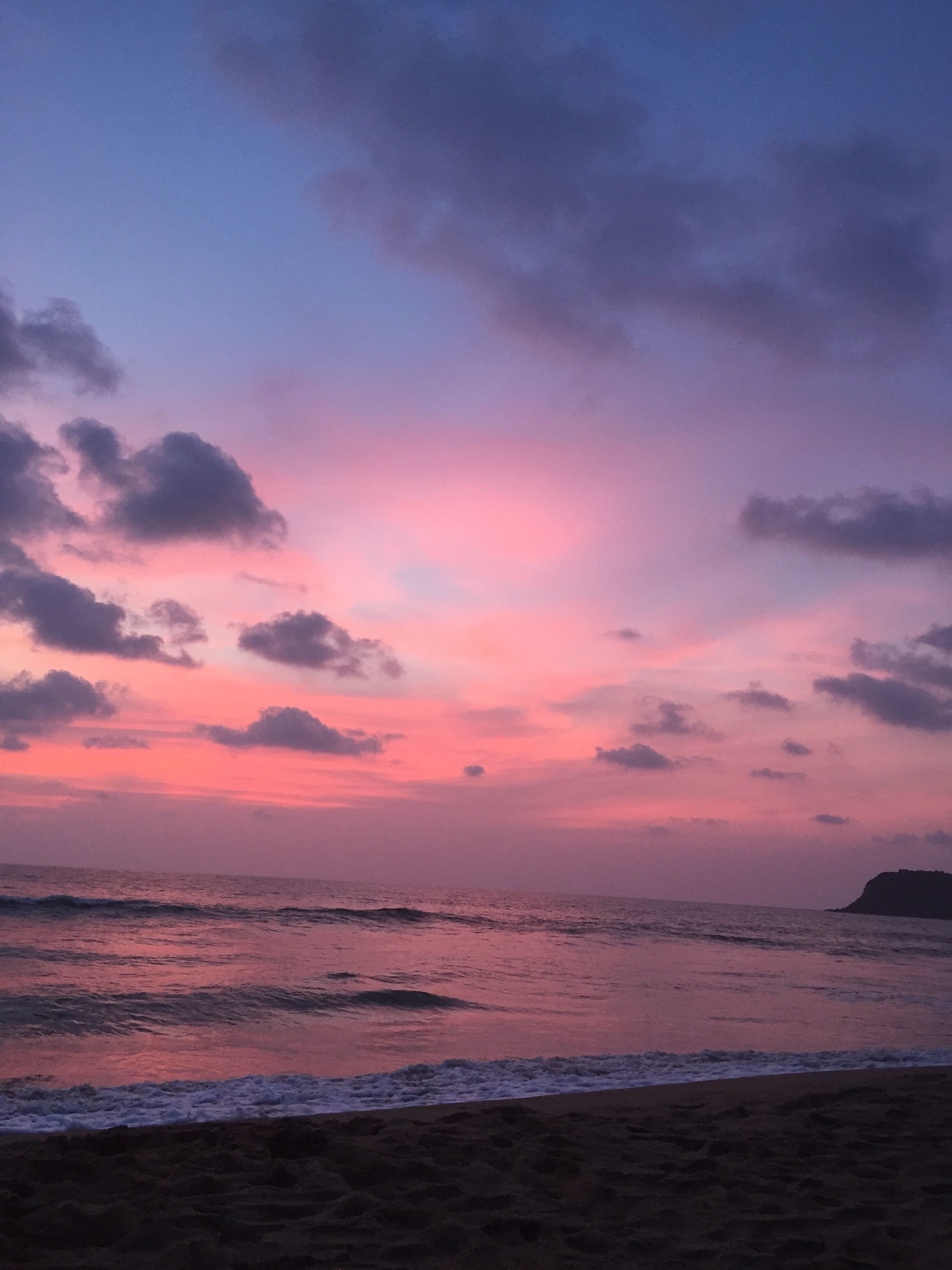 A beach at sunset with pink and purple clouds, waves, and a distant landform.