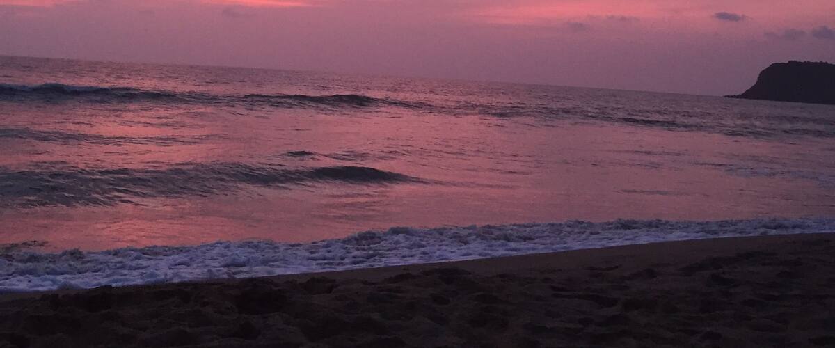 A beach at sunset with pink and purple clouds, waves, and a distant landform.