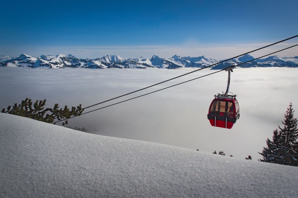 A red cable car ascending a snowy mountain slope with snow-covered peaks in the background.