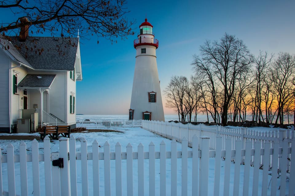 Un phare blanc avec un sommet rouge, une clôture blanche à piquets et une maison avec des volets verts.