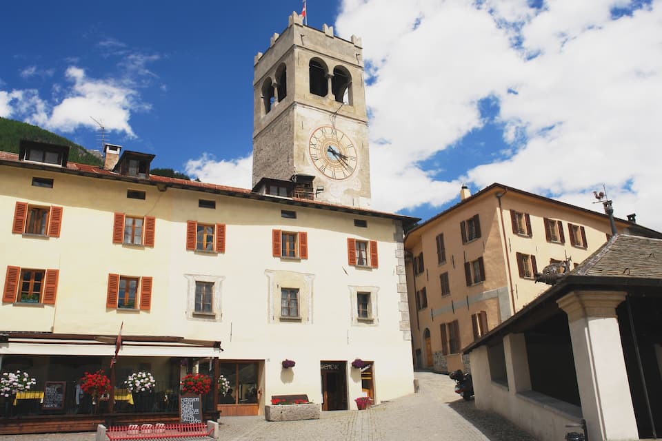 Una plaza de pueblo con una torre del reloj, edificios con contraventanas y calles adoquinadas.