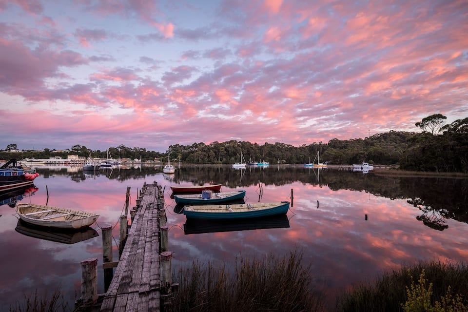 Een rustig meer bij zonsondergang, met bootjes afgemeerd aan de steiger en een bebost landschap op de achtergrond.