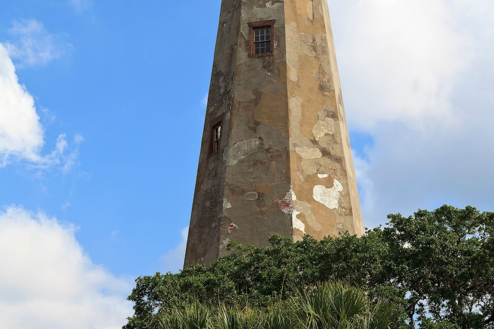 Un phare imposant, aux murs érodés par les éléments et coiffé d’un dôme, entouré d’une végétation luxuriante et se dressant sous un ciel bleu azur.