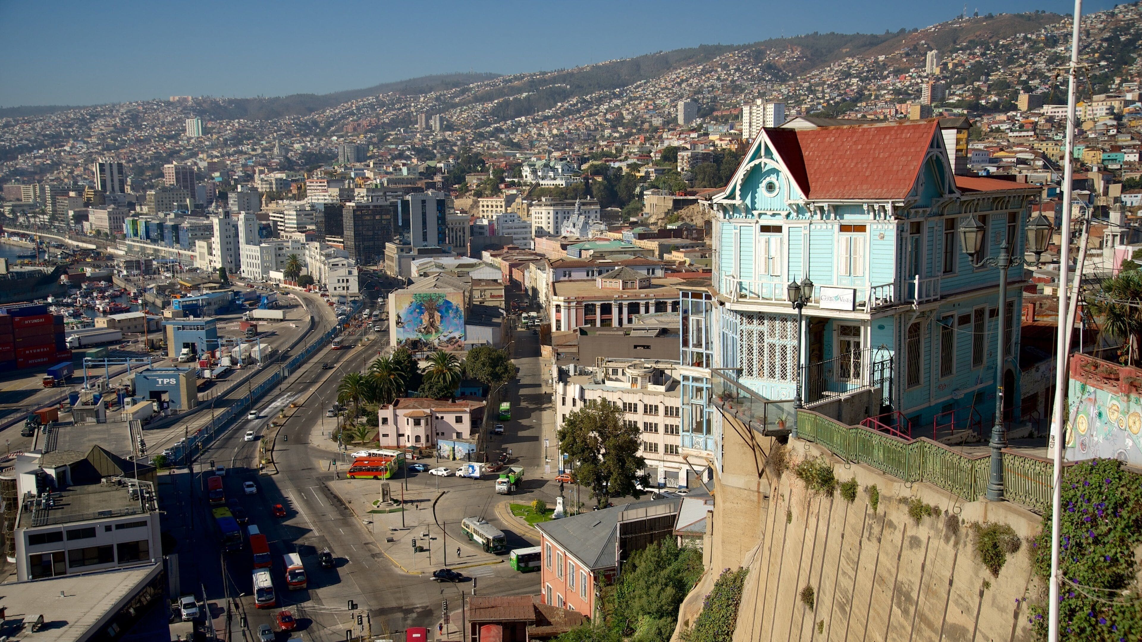 A cityscape with a prominent blue building with a red roof, a busy intersection with buses and cars, and a hilly background.