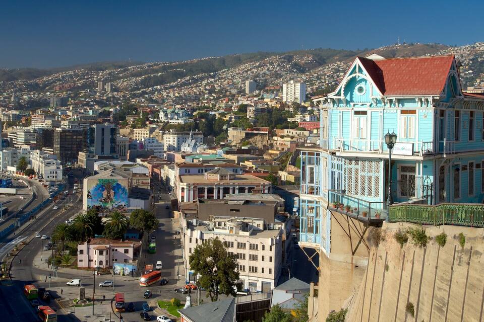 Un paisaje urbano con un edificio azul destacado de techo rojo, una calle concurrida con coches y autobuses, y un fondo montañoso.