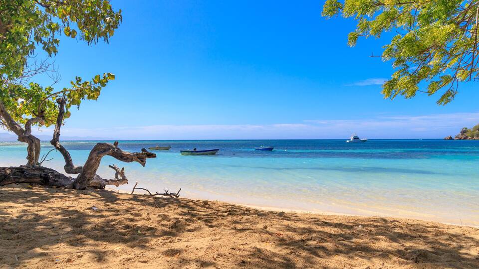 A beach with clear turquoise water, boats, and a distant island.