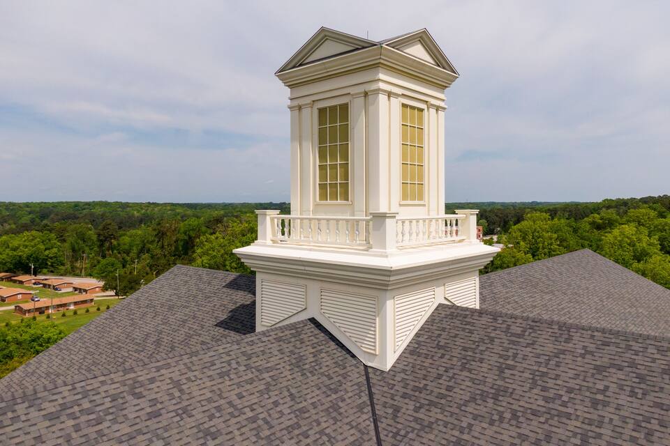 A white tower house with a balcony on a roof, surrounded by trees and a distant landscape.