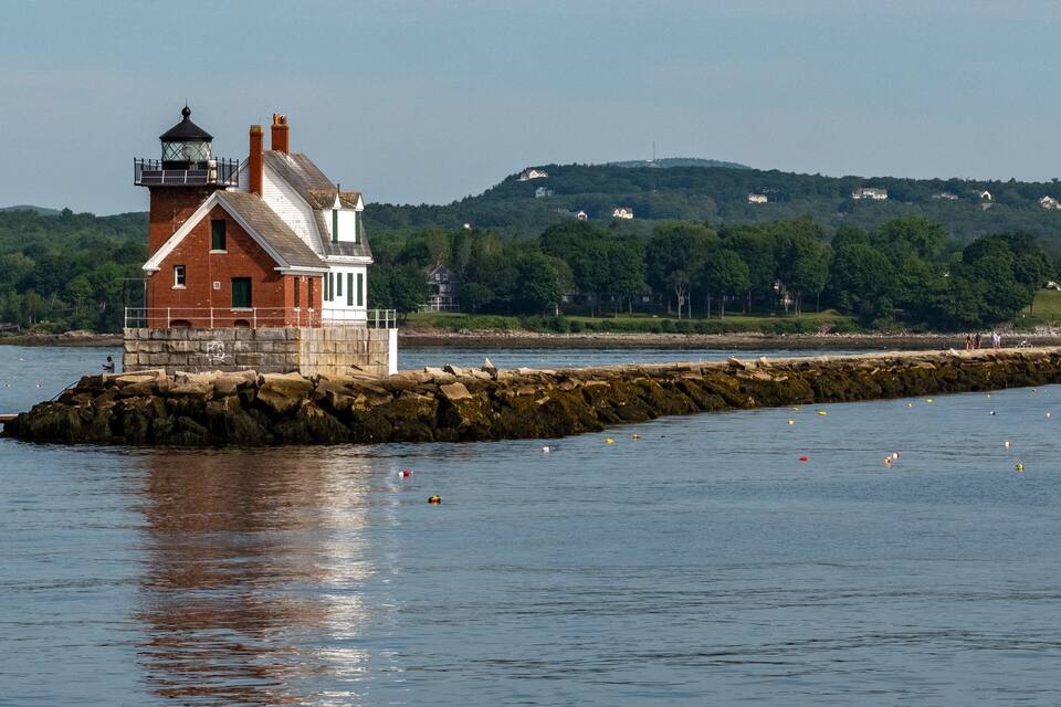 A lighthouse on a rocky breakwater with a forested hill in the background.