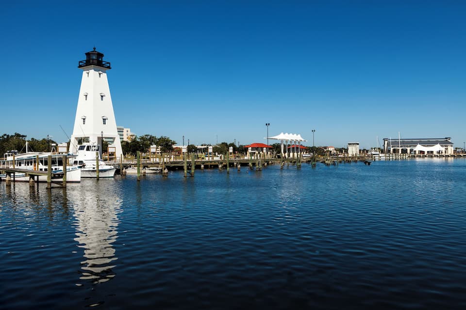 Um farol branco, uma marina com barcos e um edifício grande, de cor branca, com telhado vermelho.