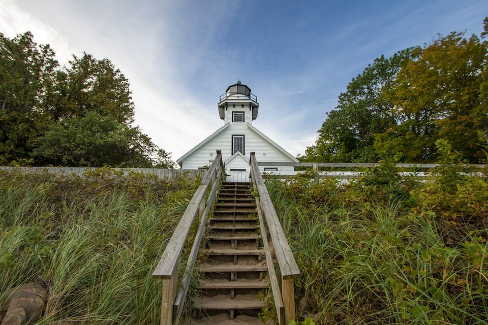 A white lighthouse with a wooden staircase leading up to it, surrounded by tall grass and trees.