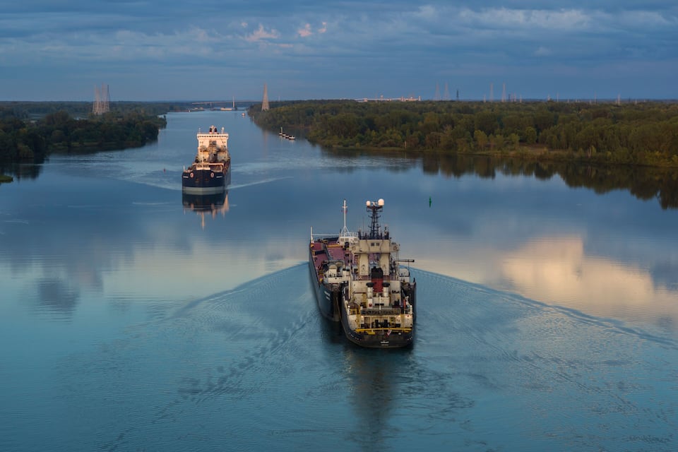 To store skibe på en stille flod med industrielle bygninger i baggrunden.