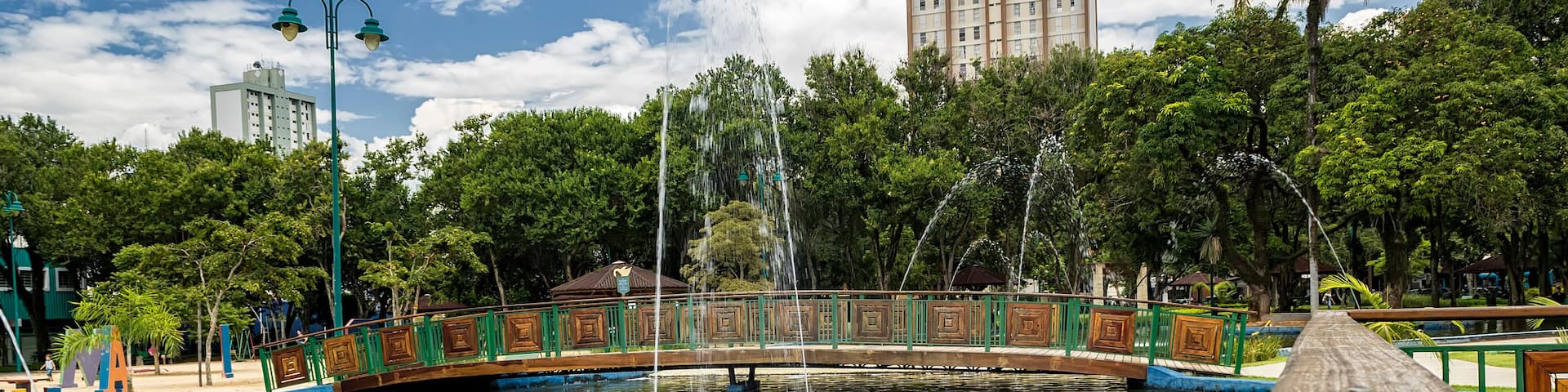 A park with a fountain, a wooden bridge, and a tall building in the background.