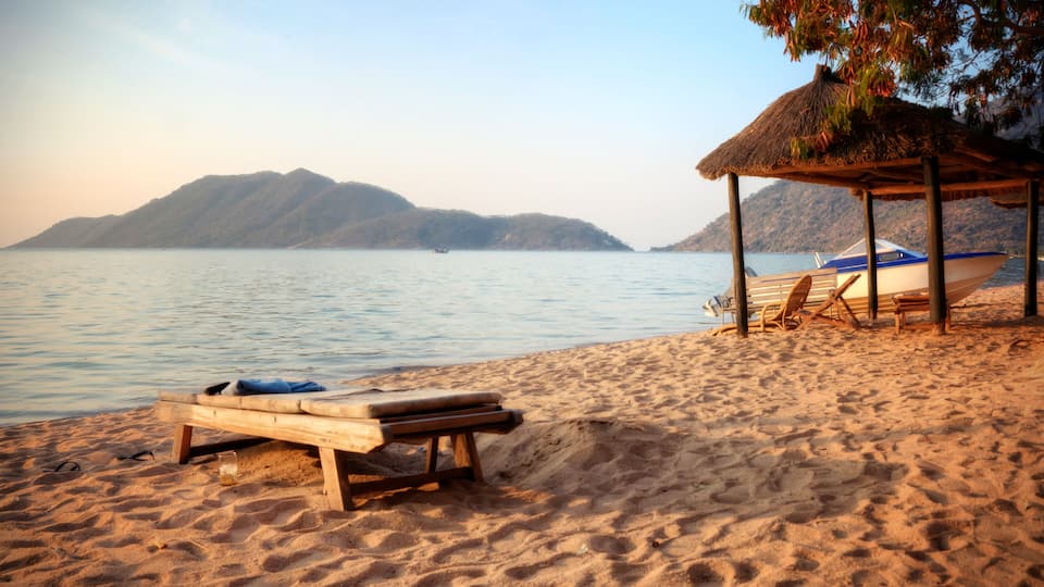A beach with a wooden lounge chair, a thatched-roof hut, and a boat on the sand.