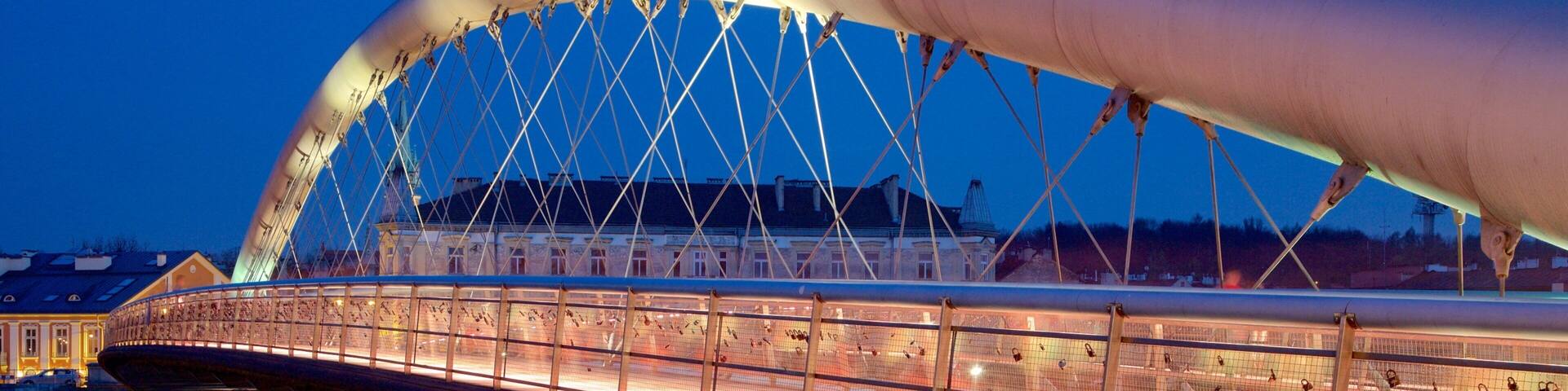 Un puente moderno con diseño curvo, iluminado por la noche, que cruza un río tranquilo con un paisaje urbano de fondo.