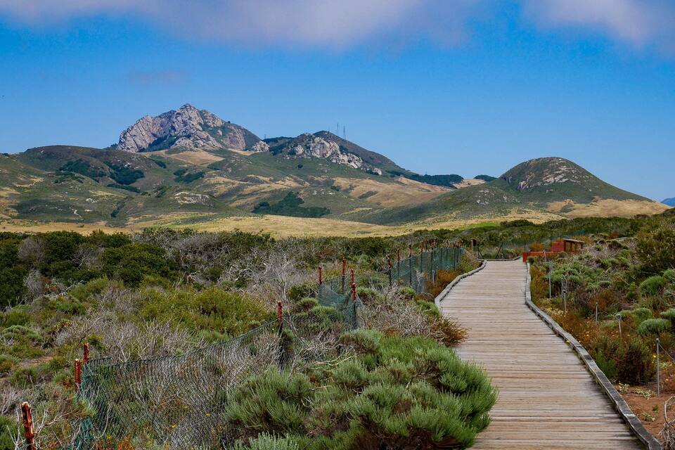 Uma passarela de madeira serpenteia por uma paisagem montanhosa, com afloramentos rochosos e vegetação esparsa.