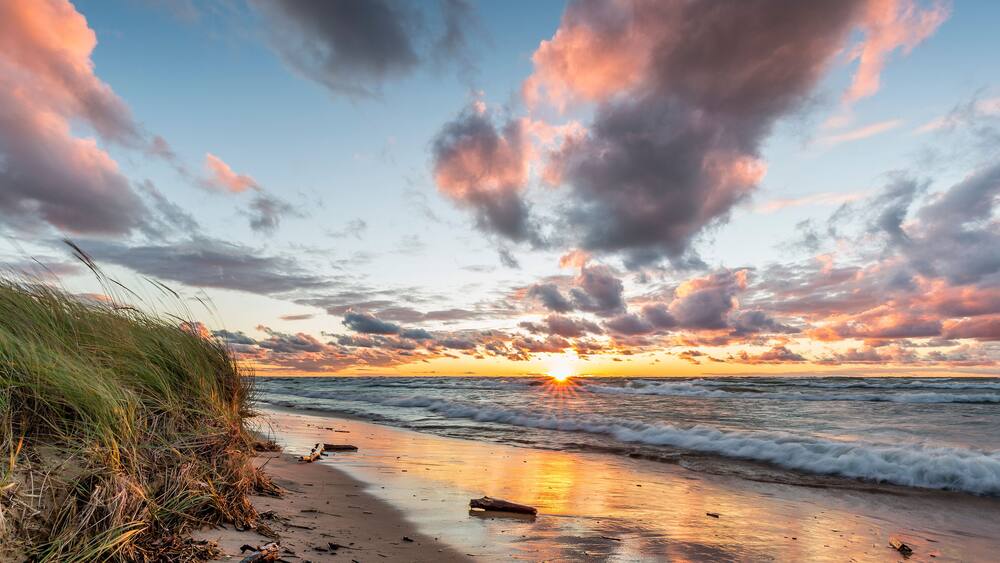 A beach at sunset with waves, grass, and clouds.