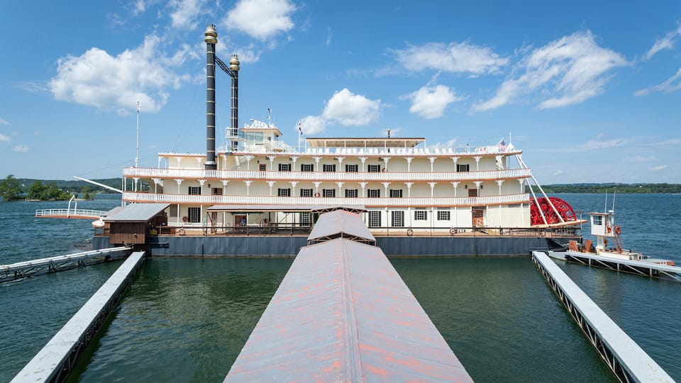 A large paddle steamer docked at a pier with a prominent smokestack and a red paddle wheel.