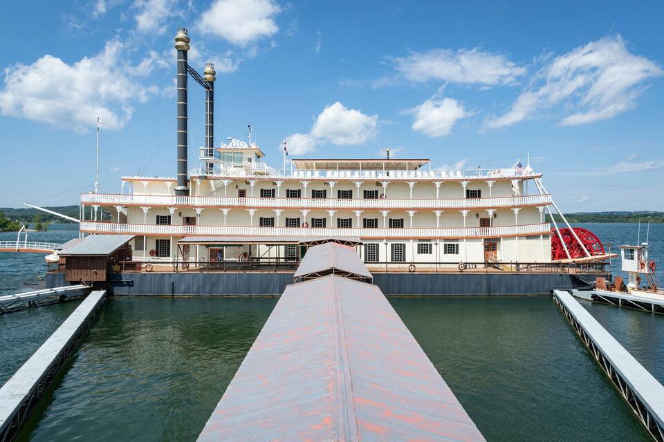 A large paddle steamer docked at a pier with a prominent smokestack and a red paddle wheel.