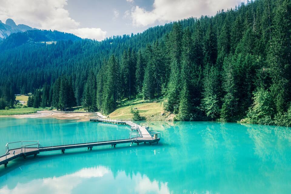Un lac turquoise, avec une jetée en bois qui s’avance sur ses eaux, entouré d’une forêt dense et de montagnes.