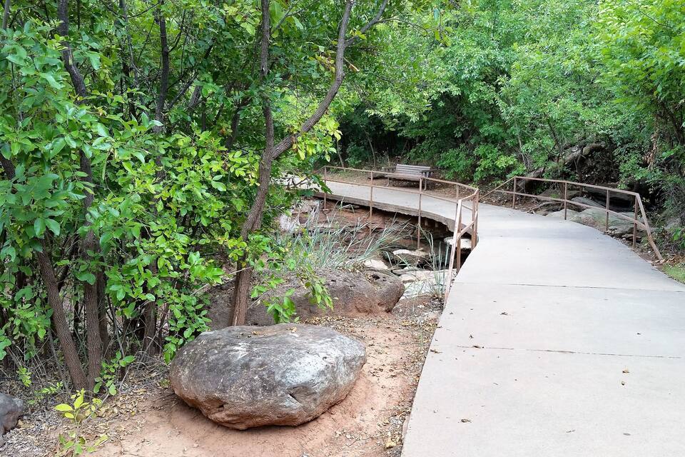 A concrete pathway, a wooden bridge, and a large rock in a forested area.