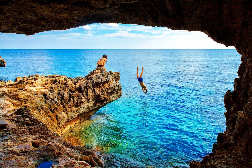 A rocky coastline with a cave entrance, clear blue sea, and two individuals engaged in water activities.