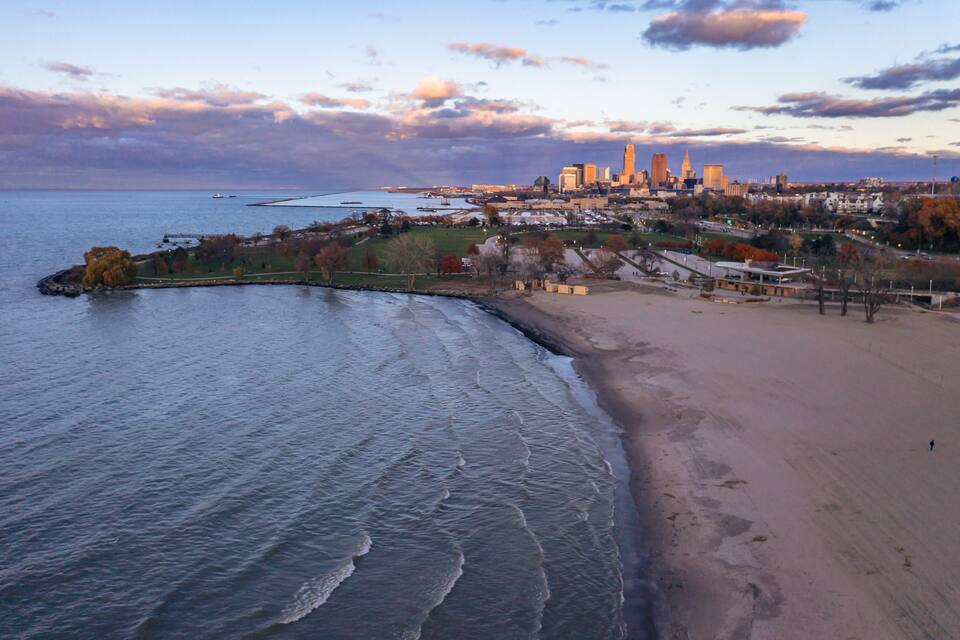A coastal cityscape with a sandy beach, a curved breakwater, and a skyline with prominent buildings.