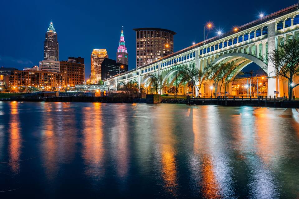 A city skyline with illuminated buildings, a bridge, and a river reflecting the lights.