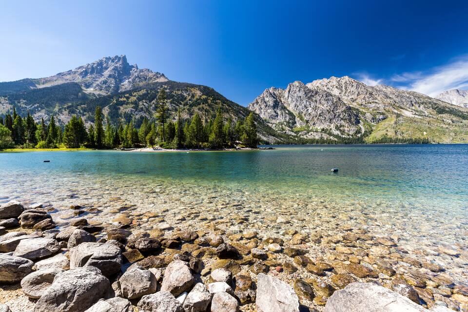 A clear lake with rocky shore, surrounded by mountains and trees.