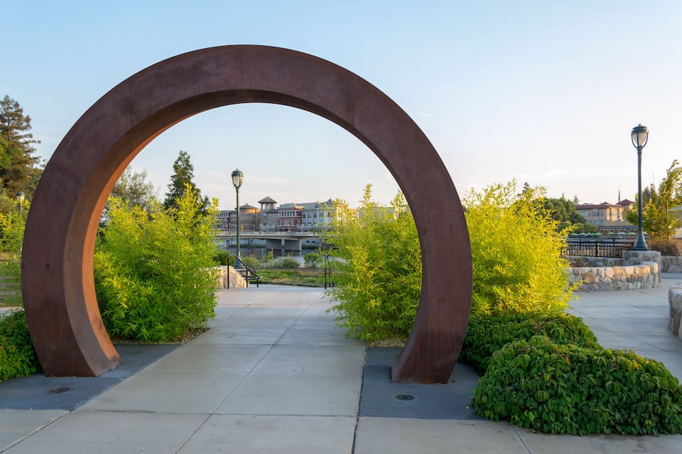 A large metal arch structure in a park with greenery and buildings in the background.