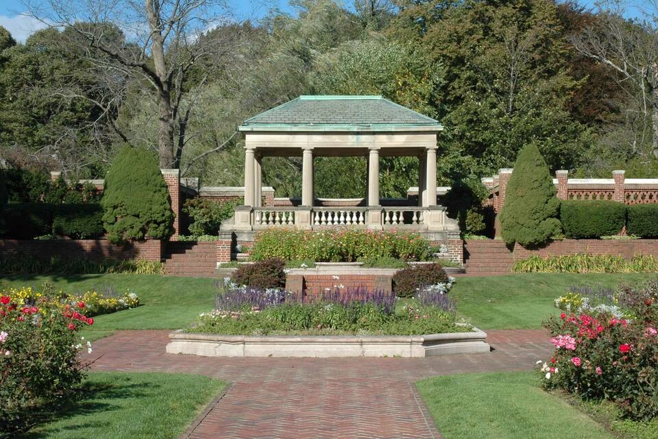 A gazebo with a green roof, surrounded by a well-manicured garden with various flowers and shrubs.