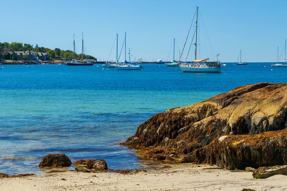 A coastal scene with clear blue water, rocky shoreline, and several sailboats anchored in the distance.