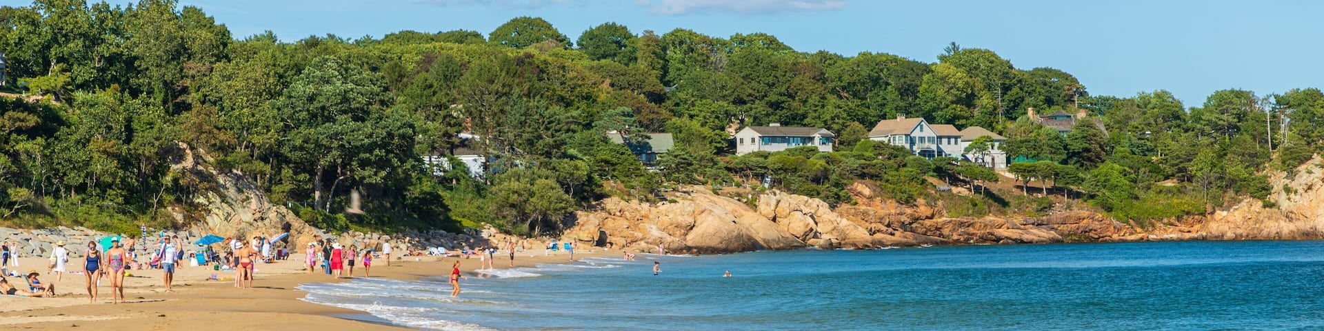 A sandy beach with people, a rocky shoreline, and houses in the background.