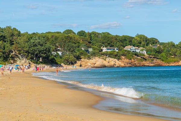 A sandy beach with people, a rocky shoreline, and houses in the background.