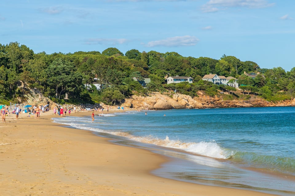 A sandy beach with people, a rocky shoreline, and houses in the background.