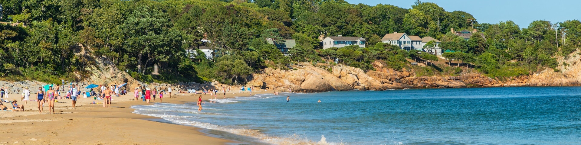 Uma praia arenosa com pessoas, uma linha costeira rochosa e casas ao fundo.