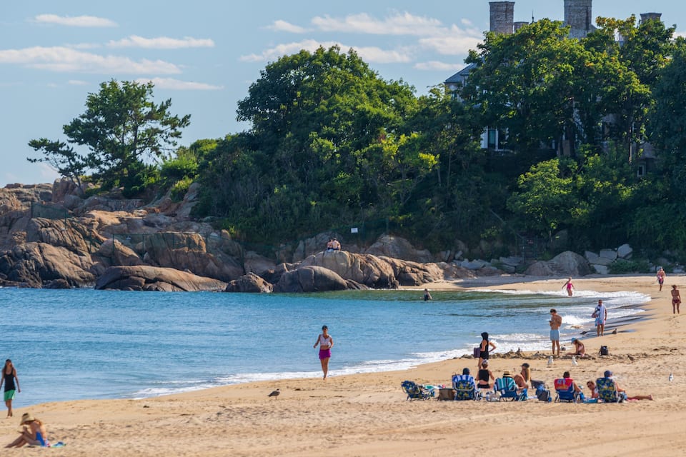 A beach with people sunbathing and swimming, surrounded by large rocks and trees.