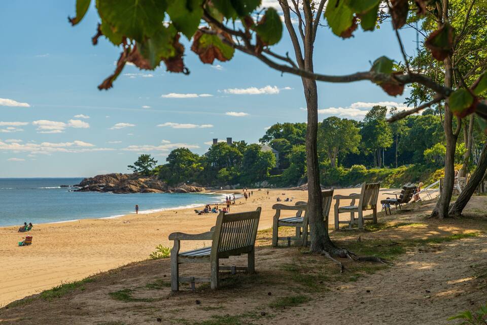 Platja de sorra, aigua i poques persones. Hi ha arbres i una zona rocosa al fons.