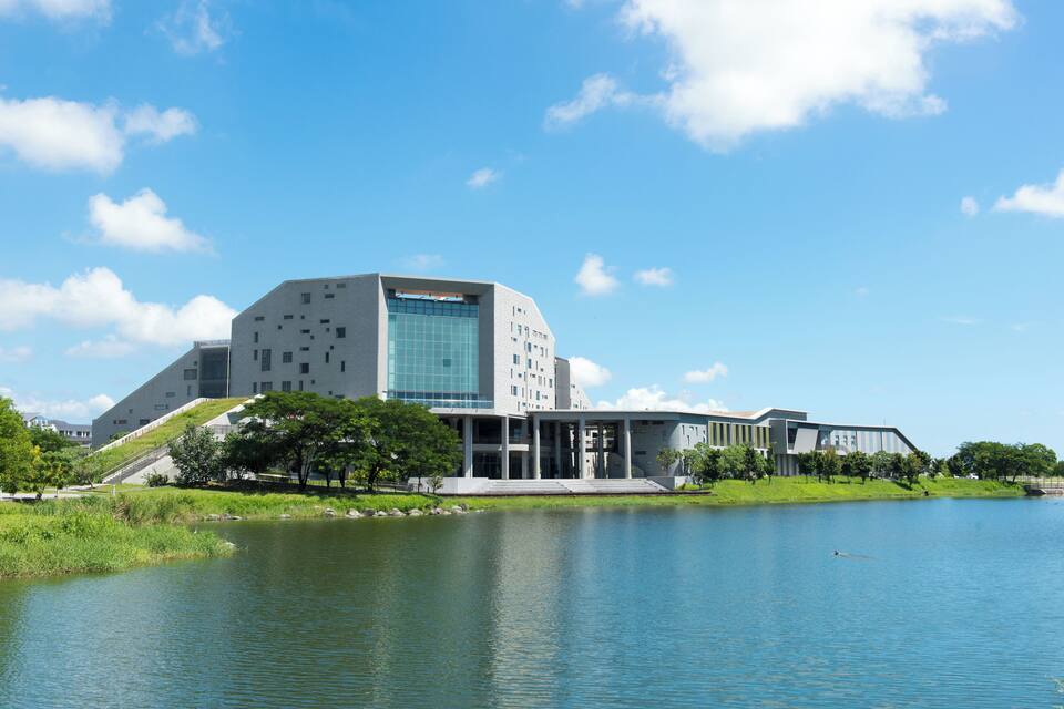A modern building with a glass facade near a calm lake, surrounded by greenery and a clear blue sky.