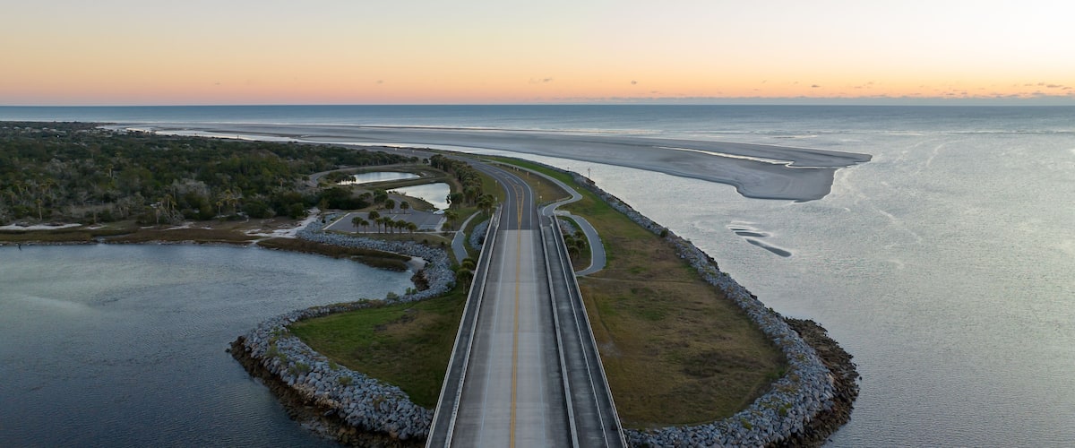 A coastal road with a bridge crossing over water, surrounded by greenery and a clear sky.