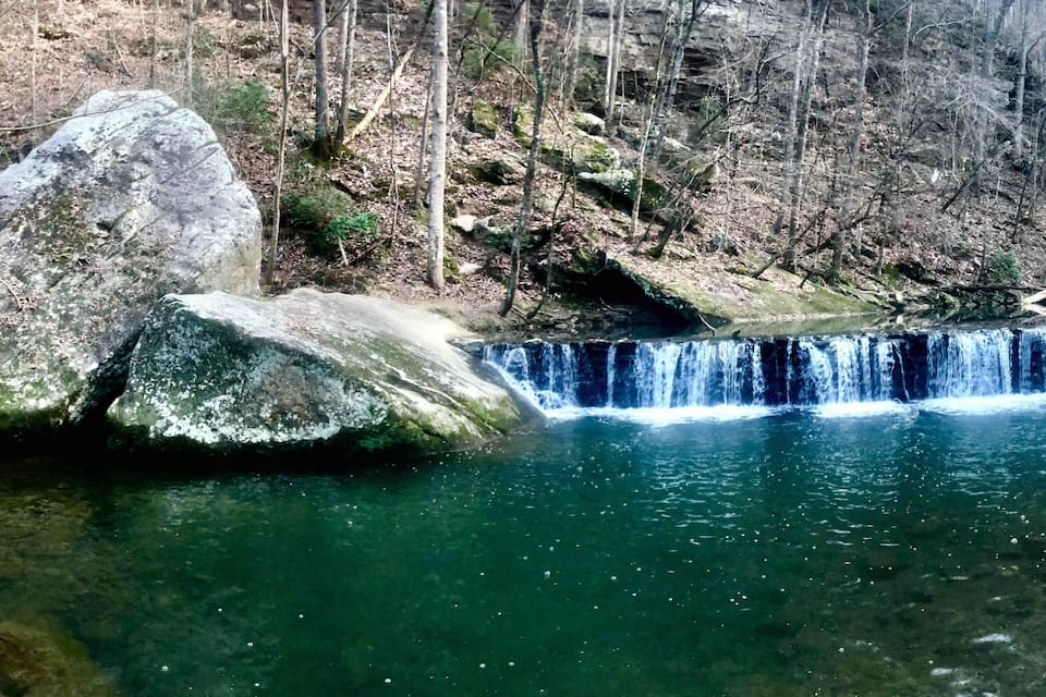 A waterfall cascading over large rocks into a clear pool surrounded by a forested area.