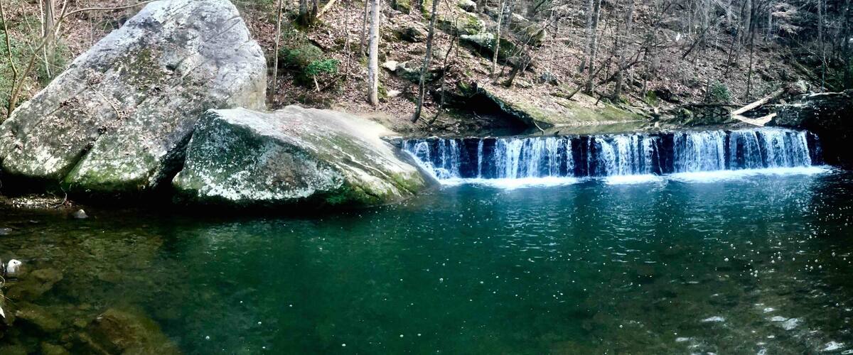 A waterfall cascading over large rocks into a clear pool surrounded by a forested area.
