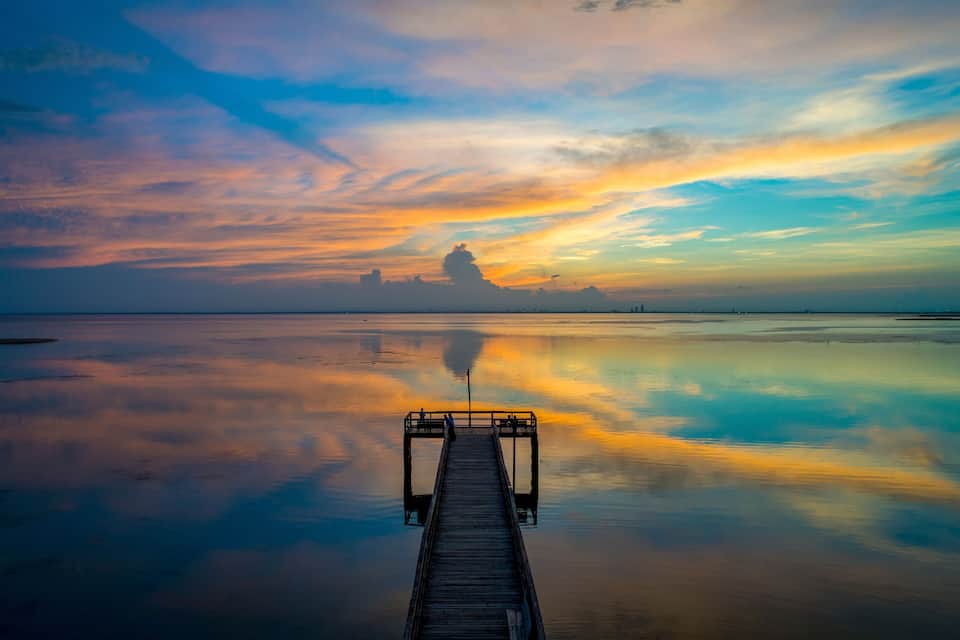 A wooden pier extending into calm waters at sunset with a colorful sky reflected on the water.