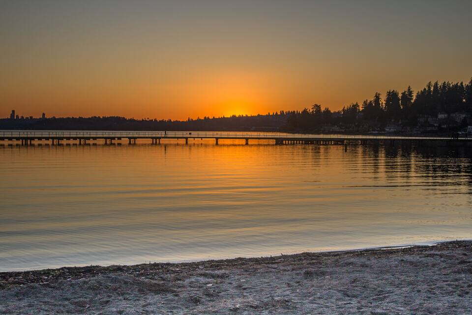 A sunset over a calm lake with a bridge and forested shoreline.