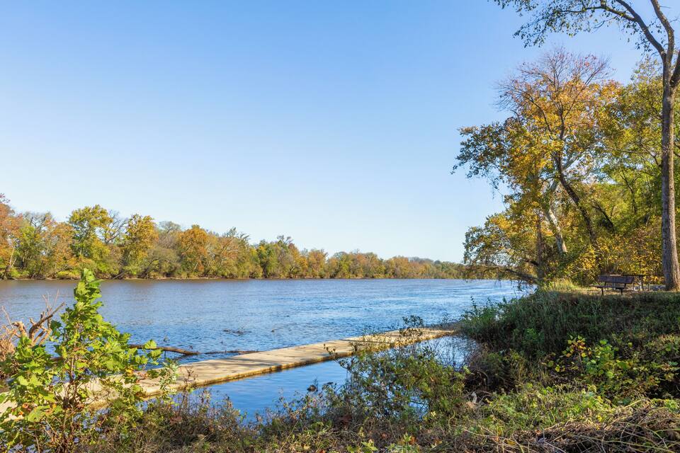 A calm river with a wooden dock extending into the water, surrounded by trees with autumn foliage.