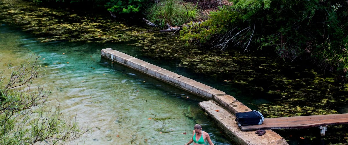 Une femme vêtue d’un maillot de bain vert se tient dans une rivière aux eaux claires, entourée d’une végétation luxuriante et d’un pont en bois.