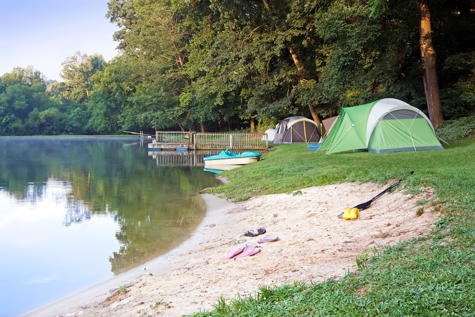 A lakeside campsite with a green tent, a canoe, and a pair of sandals on the sand.