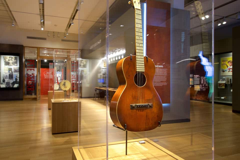 A guitar on display in a museum with wooden floors and glass cases.