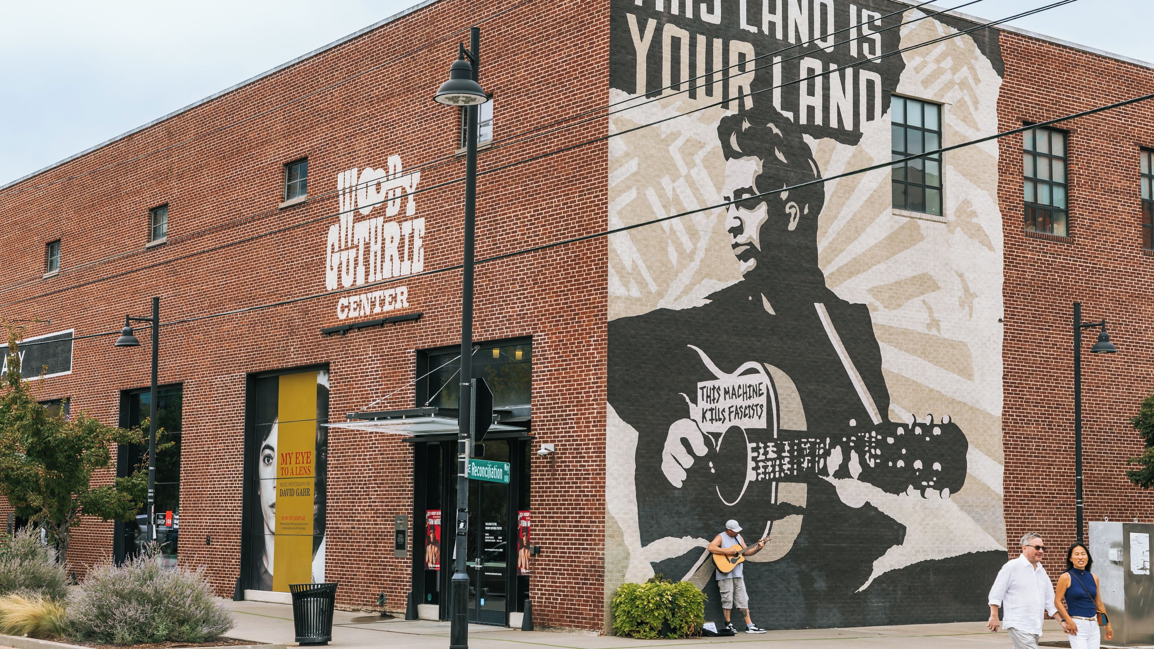Edifício de tijolos com um mural de um homem segurando um violão e uma placa com a inscrição 'THIS MACHINE KILLS FASCISTS'.