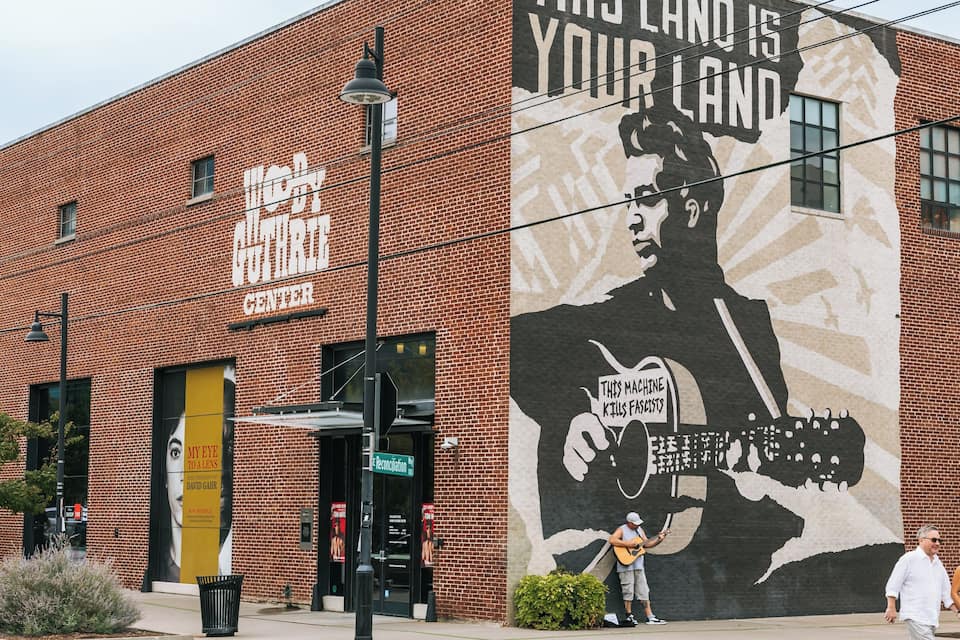 A brick building with a mural of a man holding a guitar and a sign that reads 'THIS MACHINE KILLS FASCISTS.'